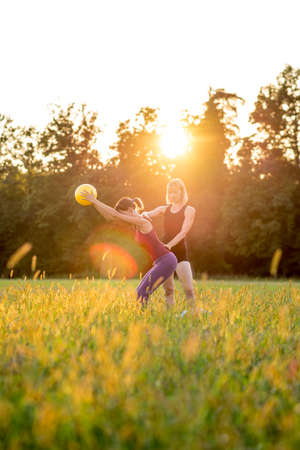 Two fit young women working out in a green field exercising with a gym ball doing pilates backlit by the glow of the rising sun over woodland trees.の写真素材