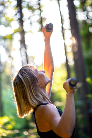 Young blond woman working out outdoors in woodland lifting weights extending the dumbbells to the glow of the sun through the trees with a happy smile.の写真素材