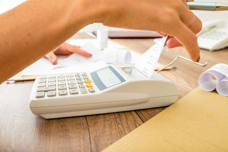 Bookkeeper doing calculations on an adding machine checking the totals against those in the journals, close up of her hands and the machine on a wooden desk.の写真素材