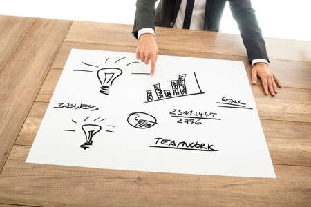 Teamwork concept in business with a businessman seated at a desk pointing to a hand drawn doodle pictogram of light bulbs, charts and figures depicting bright ideas, brainstorming and success.の写真素材