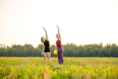 Two young fit women doing pilates exercise lifting one arm in the air and leaning with the upper body to the side outside in a beautiful nature.の写真素材