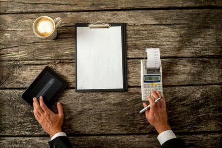 Hand of a Businessman on a Rustic Wooden Table with Tablet Computer, Calculator, Cup of Coffee and White Paper with Copy Space on a Clipboard.の写真素材