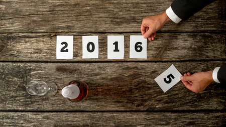 Businessman celebrating year 2015 anticipating coming year 2016 as he changes number 5 for number 6 on 2016 date on papers lying on rustic wooden desk with champagne bottle and glass on the side.の写真素材
