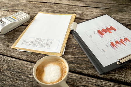 Tax and finance concept - paperwork with graphs, charts, data and numbers, adding machine and a cup of fresh hot cappuccino on a textured rustic wooden desk.の写真素材