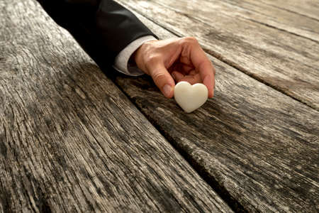Male hand in elegant suit holding heart made of white marble against a textured wooden background. Conceptual of love, romance and proposal.の写真素材
