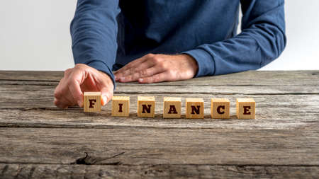 Front view of man in blue shirt assembling the word Finance with seven wooden cubes with letters on them on a rustic wooden desk.の写真素材