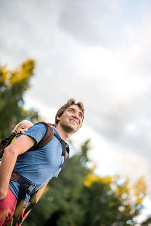 Young father carrying his child in a carrier on his back while smiling with contentment looking towards the sky in a beautiful nature.の写真素材