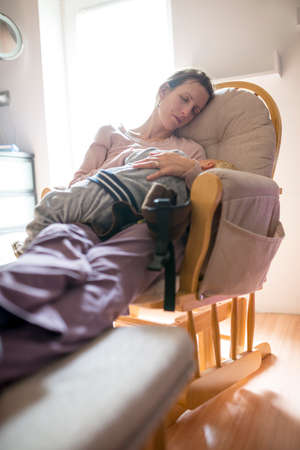 Woman resting on a comfortable chair indoors during the day with her feet up while cradling her child on her lap.の写真素材