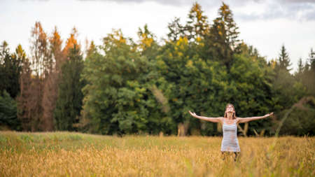 Young woman standing in the middle of autumn meadow with high golden grass looking up towards the sky with her arms spread widely as she enjoys fresh air and beauty of nature.の写真素材