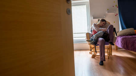 Young mother resting on a rocking chair as her toddler child sleeps in her lap.の写真素材