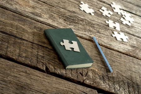 Conceptual image of knowledge, wisdom and research - a piece of puzzle lying on a closed notebook with a pencil and other puzzle pieces lying alongside on a textured wooden desk.の写真素材