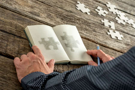 Man sketching two matching puzzle pieces in his notepad with various puzzle pieces lying on his wooden textured desk. Conceptual of education, research, solution making and innovation.の写真素材