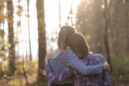 View from behind of two girlfriends or a lesbian couple standing in autumn woods leaning on each other with their arms around one another.の写真素材