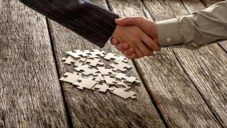 Closeup of male and female business partners shaking hands over a pile of puzzle pieces lying on a textured rustic wooden desk. Conceptual of teamwork and strategy planning.の写真素材