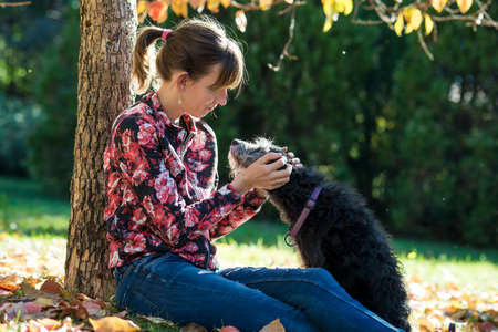 Young woman sitting under an autumn tree cuddling her black dog outside in a park.