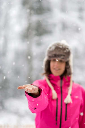 Young woman in bright pink jacket standing in a snow extending her arm in front of her to catch falling snow flakes.の写真素材