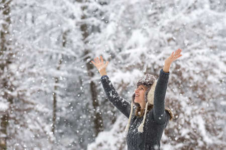 Happy young woman celebrating winter by raising her arms up in the air as she stands outside in...