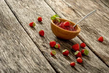 Wooden bowl full of home made strawberry marmalade with fresh ripe strawberries scattered around on textured rustic wooden desk.の写真素材