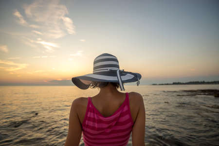Woman with striped straw hat sitting on the shore looking at the ocean under beautiful evening sky with her back to the camera.の写真素材