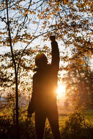 Man standing in the woods facing a setting sun with one of his arms lifted up in the air.