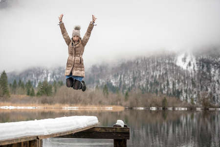 Single excited woman wearing coat and hat jumping in mid-air on snowy pier with cloud covered mountain in background.の写真素材
