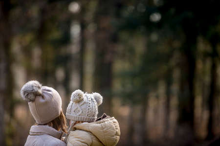 View from behind of mother holding her child both wearing warm hats outside in forested area.の写真素材