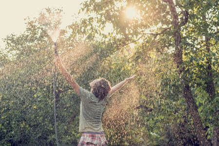 Young man outside in green summer nature holding garden hose spraying himself with arms lifted high in the air and bright sun lighting him through leaves of a tree, toned retro effect.の写真素材