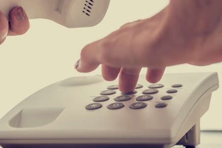 Closeup of female hand dialing a telephone number using white landline phone holding receiver with the other hand, with retro filter effect.の写真素材