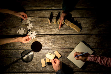 Top view of male and female business colleagues coworking on a project late at night as they take notes, stop the falling dominos, try to match puzzle pieces and make a construction of wooden pegs.の写真素材