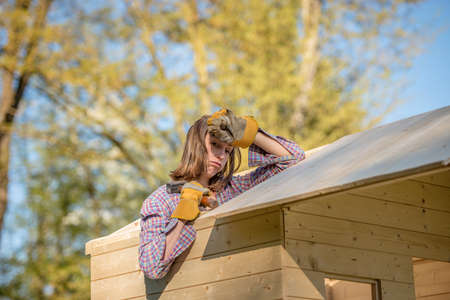 Hard working young woman with exhausted expression holding hand on forehead with hammer working on roof of tree house outdoors.の写真素材