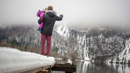 Young father in winter jacket with his back to the camera standing on the end of a snowy pier in a lake holding his toddler child pointing toward foggy mountains.の写真素材