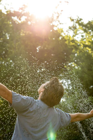 Young mam standing under a spray of water on a hot day with his arms outstretched rejoicing, leafy green tree background.の写真素材