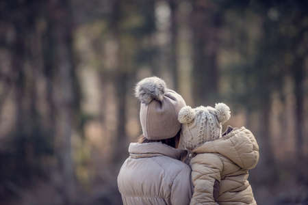 View from behind of a mother holding her child both wearing winter jackets and warm hats with blurred trees of the forest in background. With retro filter effect.の写真素材