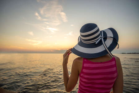 View from behind of a woman with blue and white stripped straw hat sitting on a shore looking into the distance over the ocean under an evening sky.の写真素材