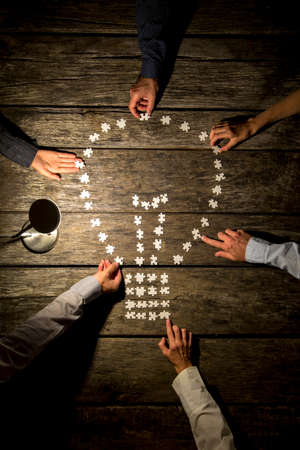 Team of six business men and women joining forces and effort to assemble a light bulb shape with blank puzzle pieces by a lit desk lamp on a textured wooden table.の写真素材