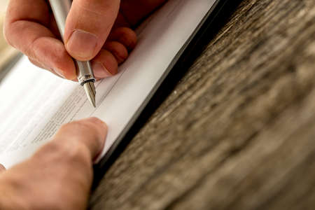 Low angle closeup view of male hand signing contract, document or application form with a pen on textured wooden desk.の写真素材
