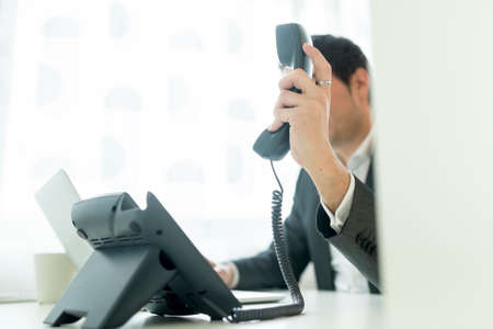 Businessman holding a telephone receiver as if about to place it on base or just has picked it up sitting at his white office desk.の写真素材