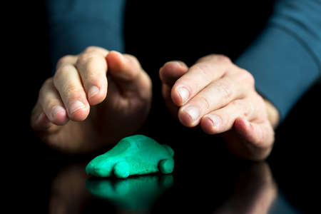Car insurance concept - male hands making protective gesture over a green car made of play dough on black desk with reflection. Over black background.の写真素材