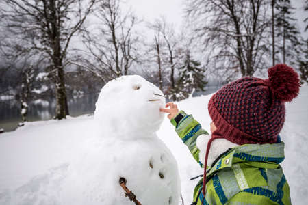 Young child wearing a warm jacket and knitted cap making a winter snowman preparing a hole for the mouth in a snowy landscape.の写真素材