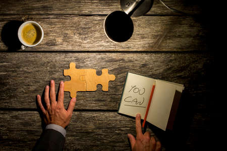 Conceptual image with inspirational message  You Can, of a man working on a puzzle on a rustic wooden table by the light of a lamp with coffee and a notebook with the handwritten words, overhead view.の写真素材