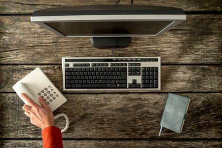 First person view of hand reaching for phone seated in front of computer keyboard, monitor and notepad.の写真素材