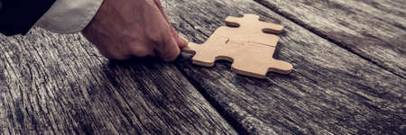 Wide crop on single hand coming from formal business suit attaching puzzle piece on old wooden table, vintage effect toned image.の写真素材