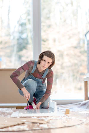 Single young adult woman in overalls working on floor with old furniture or projects in home.の写真素材
