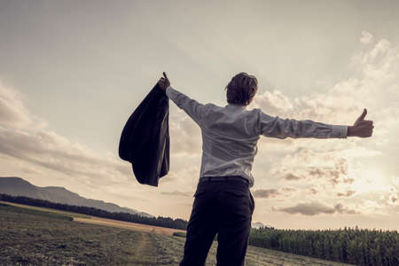Rear view of single businessman with removed jacket with his arms outspread showing thumbs up sign at the open field during sunset time.の写真素材