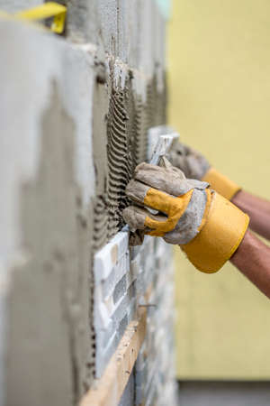 Gloved hands of worker installing ceramic wall tiles over freshly scored mortar compound.の写真素材