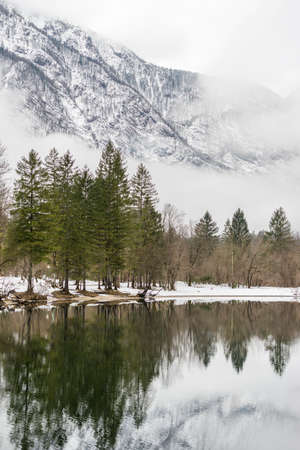 Tranquil winter lake in the mountains with the evergreen conifers reflected in the still water and snowy mountain peaks behind shrouded in wisps of cloud or mist.の写真素材