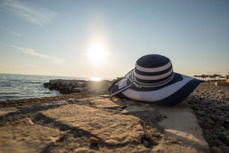 Fashionable straw sunhat at the seaside lying on a shelf of rock overlooking the ocean with the evening or morning sun low on the horizon in the background, conceptual of a summer vacation.の写真素材