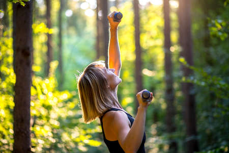 Sporty young woman working out with weights to tone and strengthen her muscles outdoors in sunlit woodland amongst the trees in a health and fitness concept.の写真素材