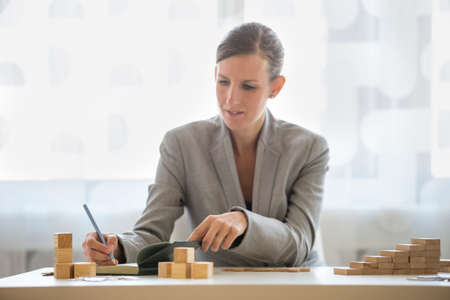 Woman in business attire writing on notepad beside stacks of wooden blocks on her desk. Includes copy space.の写真素材