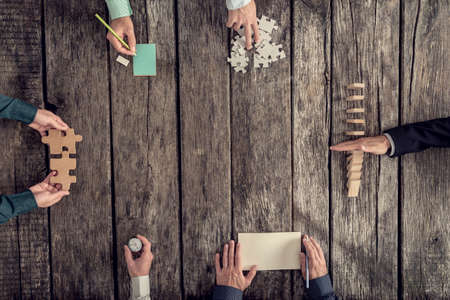 Business strategy and brainstorming concept with a team of six businessmen holding puzzle pieces, writing down ideas on paper and stopping domino effect, top view.